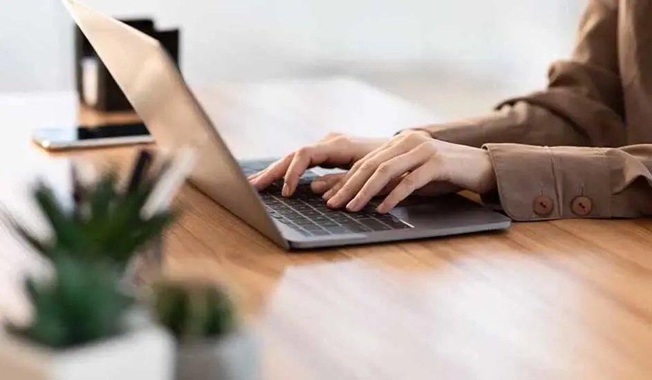 Une personne utilise un ordinateur portable sur une table en bois, avec un téléphone, un pot à crayons et une plante en arrière-plan.
