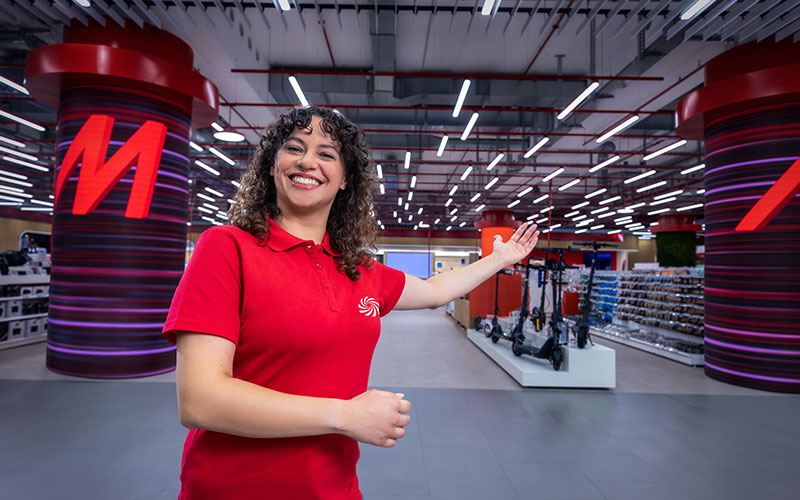 Glimlachende winkelmedewerker in rood hemd verwelkomt klanten in een grote elektronicawinkel.