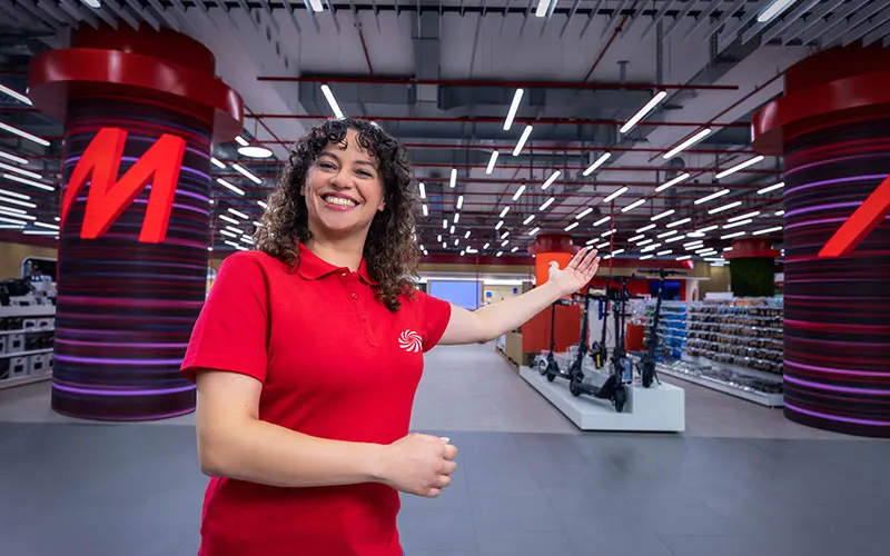 Glimlachende winkelmedewerker in rood hemd verwelkomt klanten in een grote elektronicawinkel.