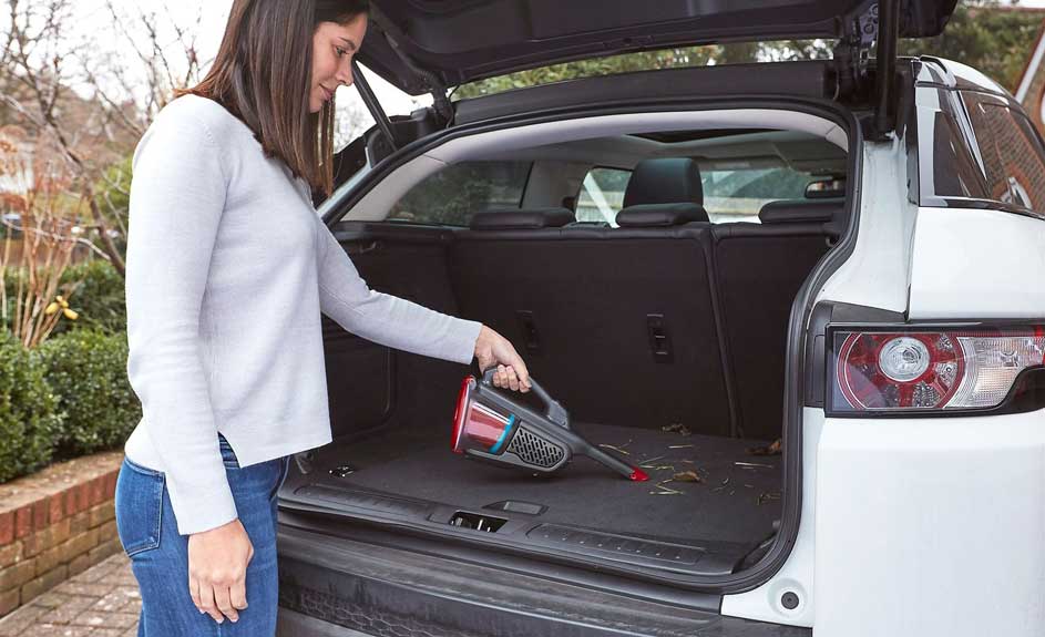 Une personne utilise un aspirateur à main pour nettoyer le coffre d'une voiture blanche. Le coffre est ouvert.