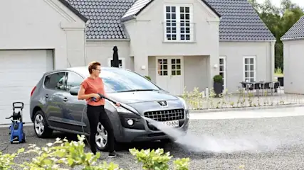 Een grijze auto wordt schoongespoten met een blauwe hogedrukreiniger voor een wit huis met grijze dakpannen.