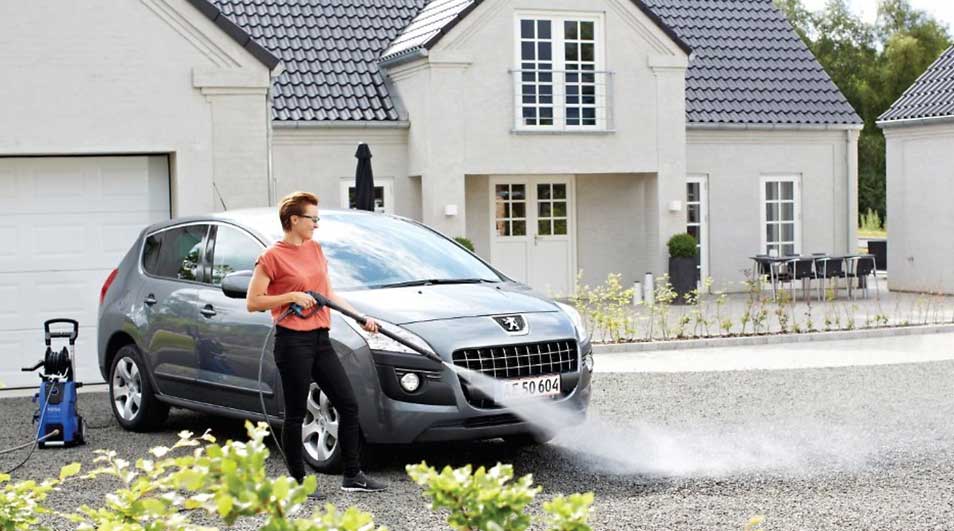 Une voiture grise est nettoyée au jet à haute pression devant une maison beige avec un toit gris.