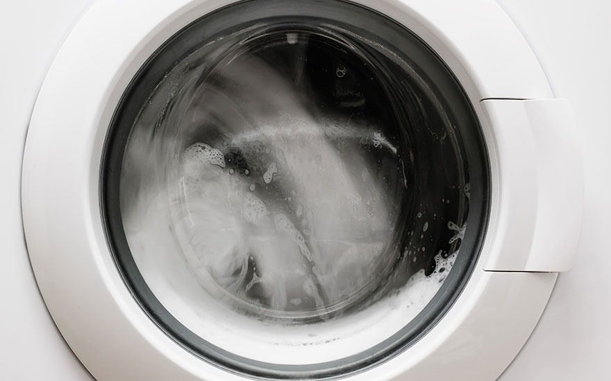 Une machine à laver blanche avec un hublot en verre montre du linge qui tourne avec de l'eau et du savon.
