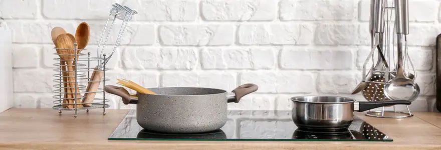 Une cuisine avec des ustensiles, une casserole grise et une autre en inox sur une plaque de cuisson en verre. Mur en briques blanches.