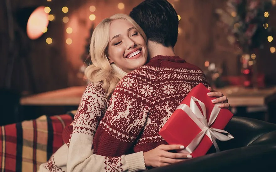 Un couple s'embrasse, portant des pulls de Noël rouges et blancs. Un cadeau rouge avec un ruban blanc est visible. Arrière-plan flou avec des lumières.