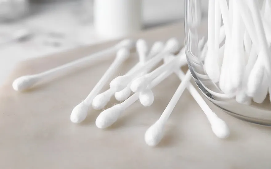 Des cotons-tiges blancs sont éparpillés près d'un pot en verre rempli de cotons-tiges sur une surface claire.