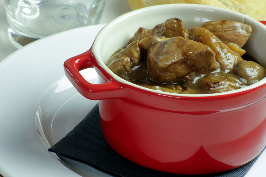 Un plat mijoté dans un bol rouge repose sur une serviette noire, sur une assiette blanche, avec un verre à eau en arrière-plan.