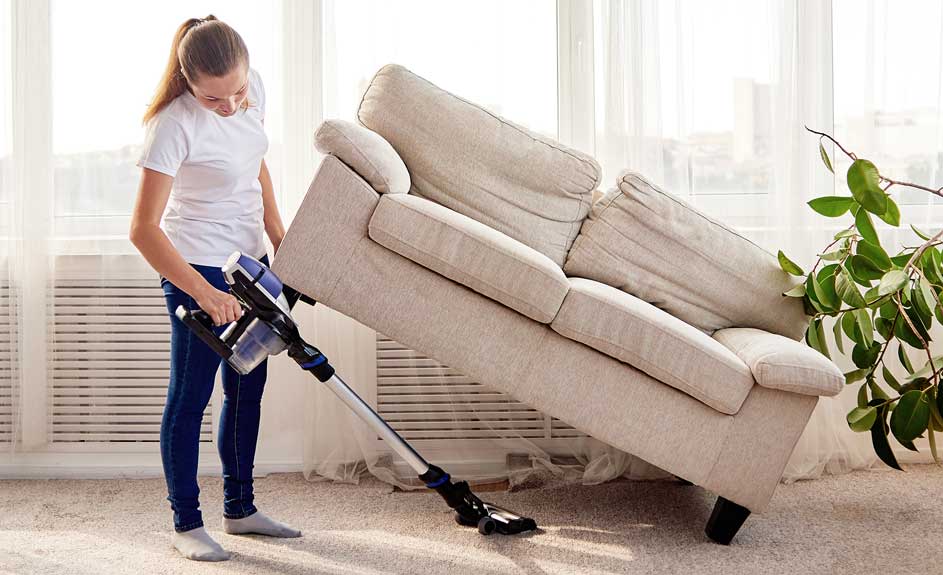 Une femme passe l'aspirateur sous un canapé beige dans une pièce lumineuse avec des rideaux blancs et une plante verte.