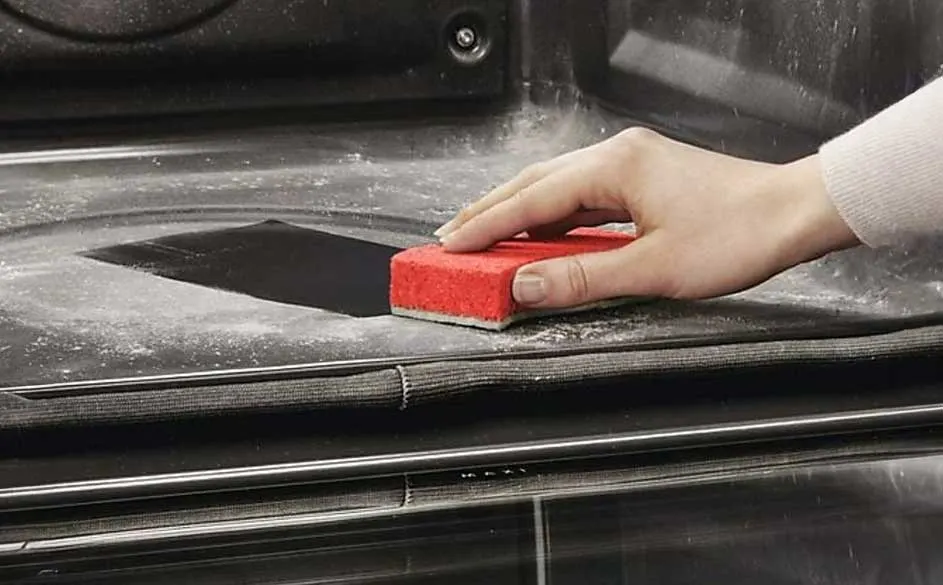 Een rode spons reinigt een zwart ovenoppervlak met een metalen handgreep, bedekt met wit poeder.