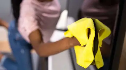 Een gele doek veegt een donker oppervlak schoon. Op de achtergrond is een roze shirt en een blauwe jeans te zien.