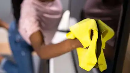 Une personne nettoie une surface sombre avec un chiffon jaune, sur fond d'objets blancs et de vêtements colorés.