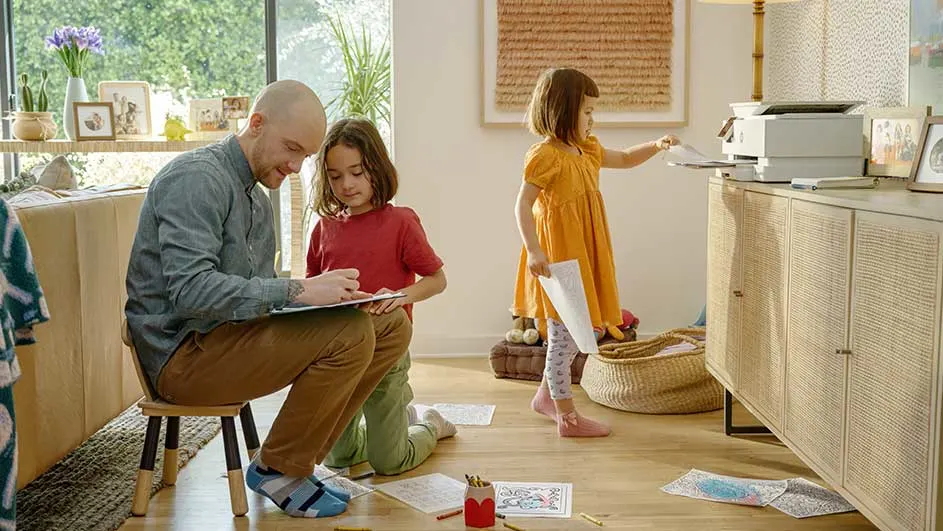 Salon clair avec un homme sur une chaise, des enfants, une imprimante sur une commode en bois. Des crayons sur le sol.