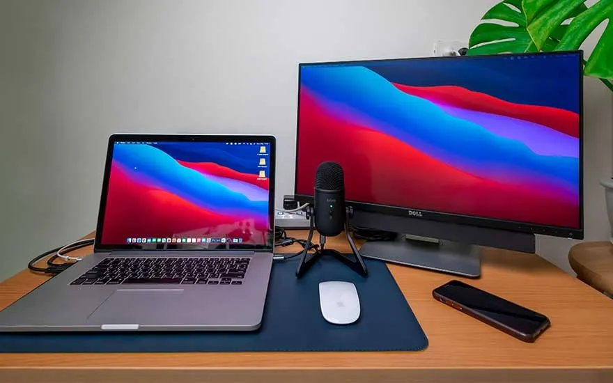 Een laptop, computerscherm, microfoon, muis en telefoon op een houten bureau met een blauwe muismat. Achtergrond is een witte muur en een groene plant.