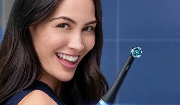 Een glimlachende vrouw met donker haar kijkt naar de camera; naast haar staat een elektrische tandenborstel met ronde borstelkop tegen blauw.