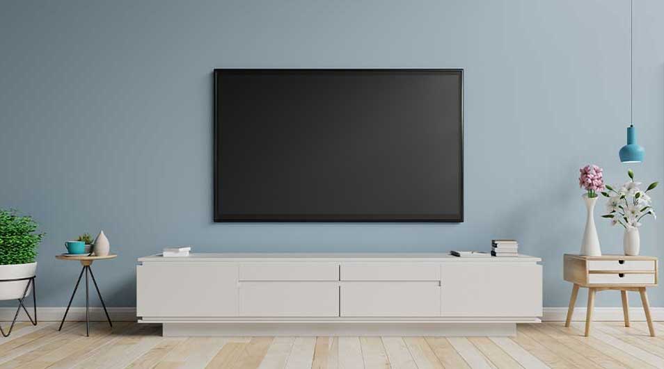 Woonkamer met blauwe muur, witte tv-meubel, houten vloer, planten, bloemen en een tv aan de muur.