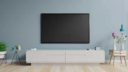 Salon avec TV noire sur mur bleu. Meuble TV blanc, tables avec plantes et fleurs. Sol en bois clair.