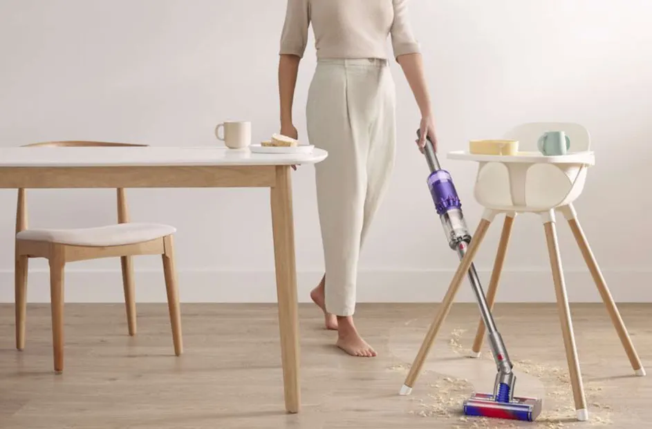 Een paarse stofzuiger zuigt kruimels op naast een houten kinderstoel. Een witte tafel en stoel staan op de achtergrond.