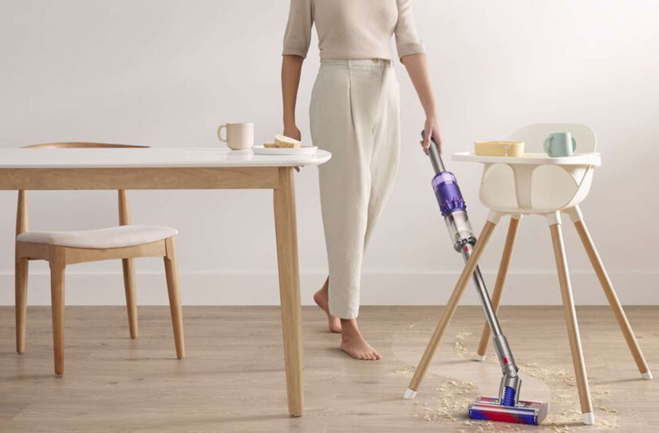 Une table blanche, une chaise, une chaise haute, et un aspirateur violet sur un plancher en bois clair.