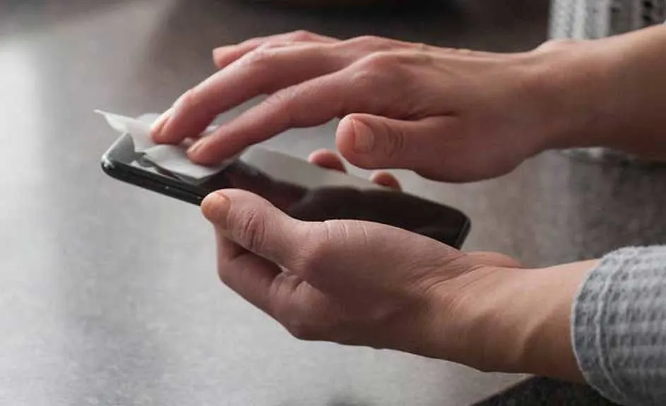 Une personne nettoie un smartphone noir avec une lingette blanche sur une surface grise, près d'un vêtement à motifs.