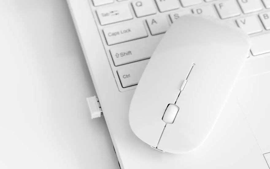 A white wireless mouse rests atop a white laptop, with visible keys and a USB receiver plugged into the side.