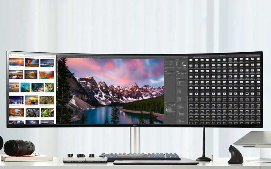 Écran incurvé ultra-large affichant des photos, avec un clavier, une souris et un appareil photo sur un bureau blanc.