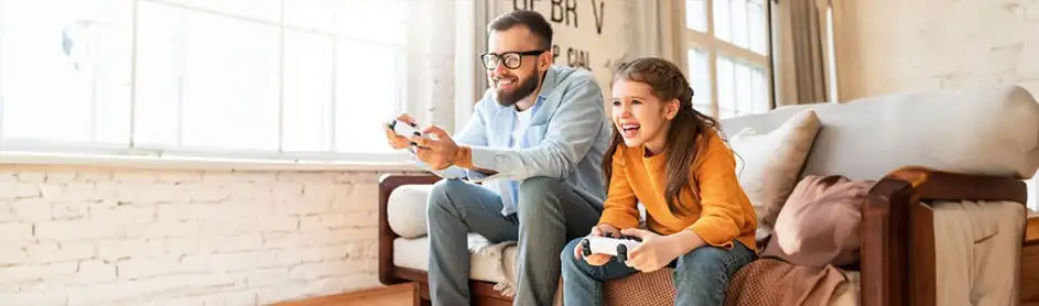 Un salon avec un canapé en bois où deux personnes jouent à des jeux vidéo avec des manettes blanches. Une fenêtre est visible à l'arrière-plan.