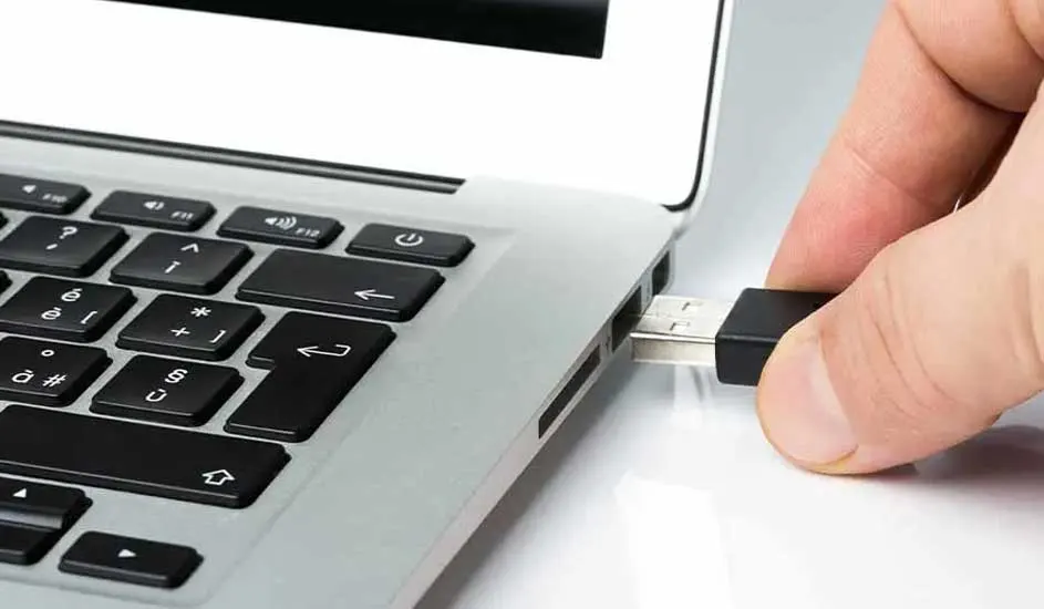 Een zilveren laptop met een zwart toetsenbord, een hand die een USB-stick in de poort steekt, op een witte achtergrond.