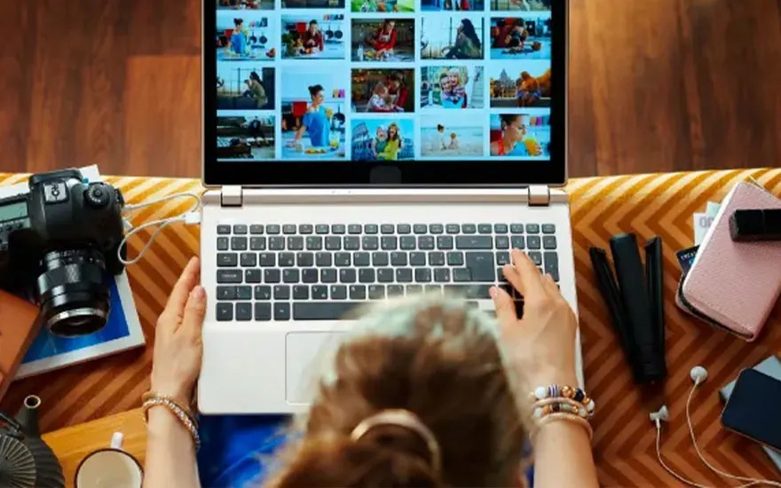 Laptop displays photos, hands type. Camera, phone, and mug on a patterned surface.