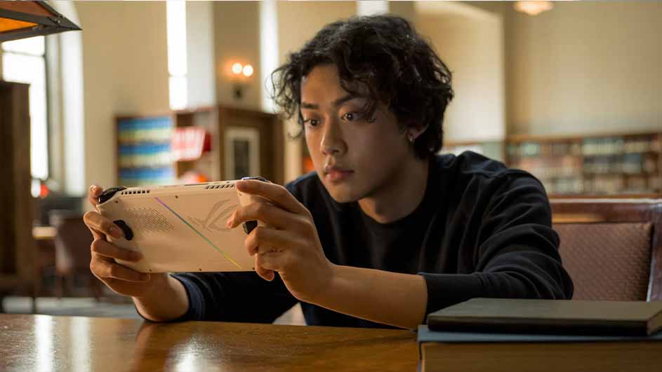 Een witte draagbare spelconsole wordt vastgehouden boven een houten tafel, met boeken en een bibliotheek op de achtergrond.