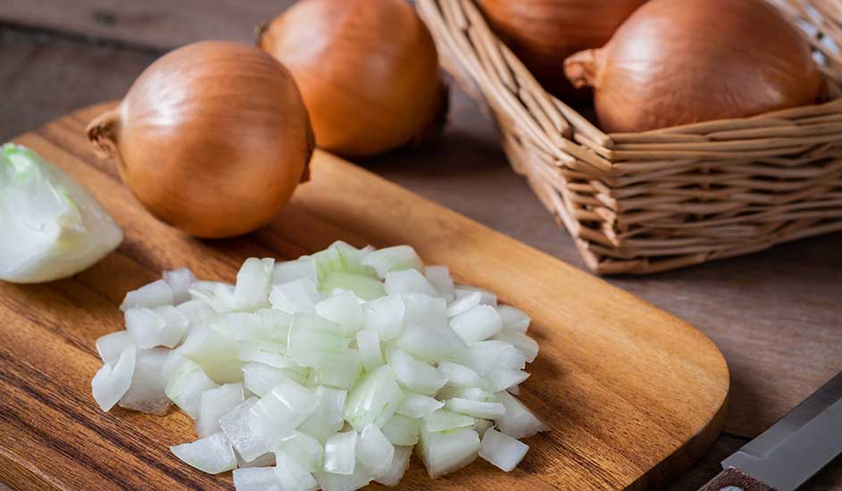 Een houten snijplank met gesneden uien en een mand met uien. De uien zijn bruin en wit van kleur.