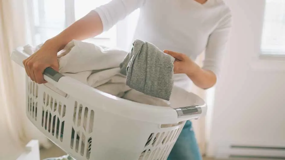 Une personne porte un panier à linge blanc rempli de vêtements près d'une fenêtre, tenant un vêtement gris.
