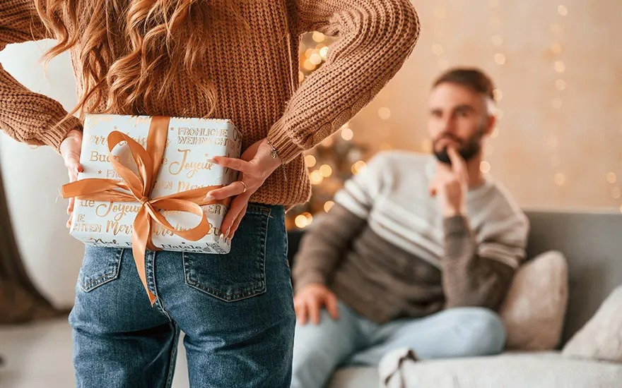 Une femme tient un cadeau derrière son dos, avec un homme assis sur un canapé en arrière-plan.