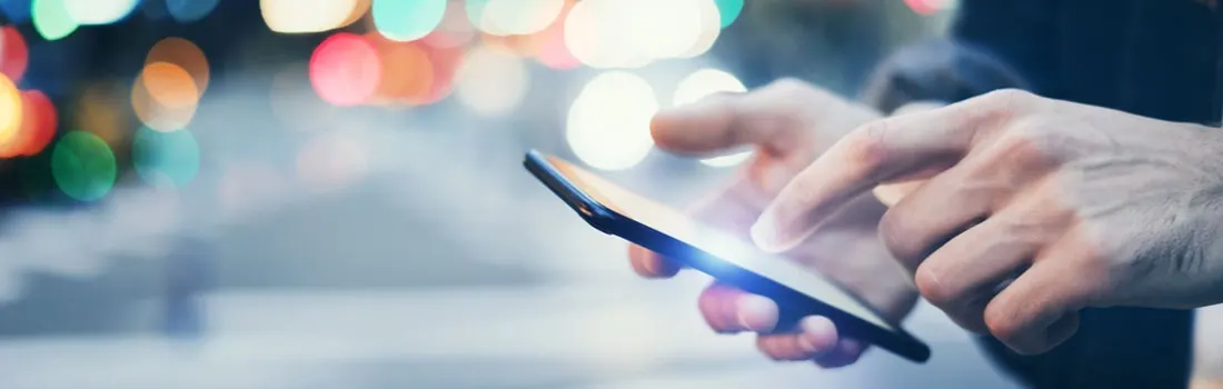 A hand holds a smartphone, with a finger touching the screen. Bokeh lights appear in the background.
