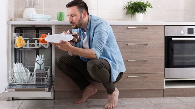 Een persoon hurkt voor een open vaatwasser in een keuken, met borden en bestek zichtbaar in de rekken.