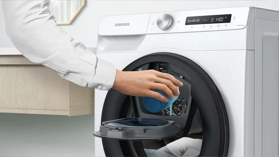 Een witte Samsung wasmachine met een open deur. Een hand giet blauwe vloeistof in een compartiment.