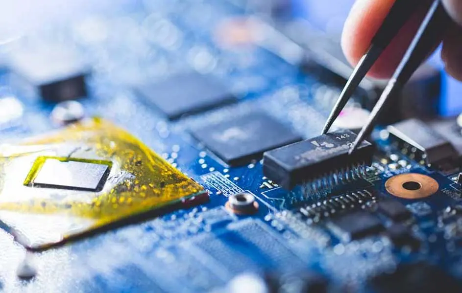 Detail van een blauwe printplaat met gouden en zwarte componenten. Een pincet houdt een zwarte chip vast.