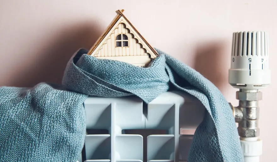 Un radiateur blanc est recouvert d'une écharpe bleue et d'une petite maison en bois. Un thermostat blanc est visible à droite.