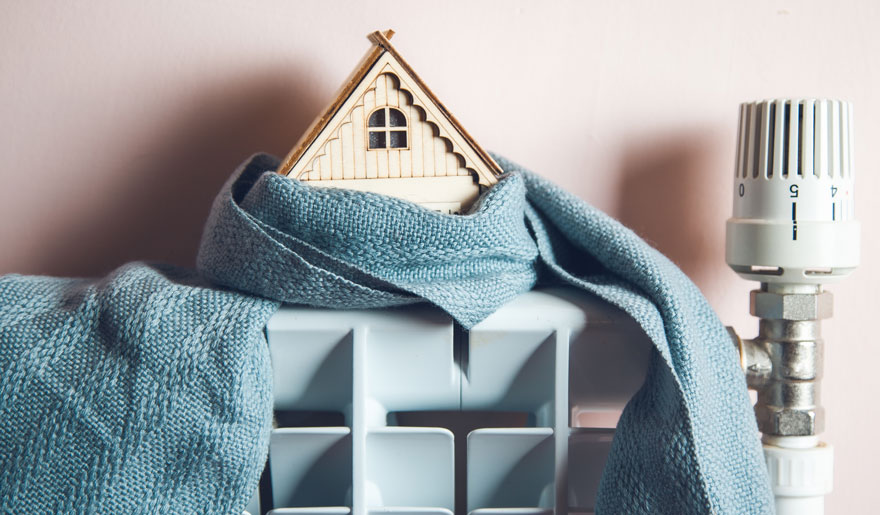 Houten huisje met blauwe sjaal op een witte radiator met thermostaat tegen een roze achtergrond.