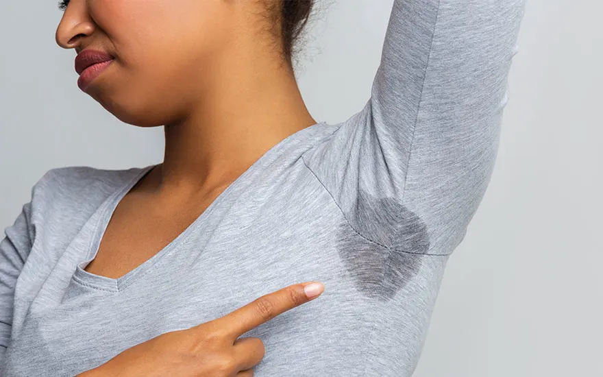 Un t-shirt gris clair présente une tache sombre sous l'aisselle, pointée du doigt sur un fond gris uni.