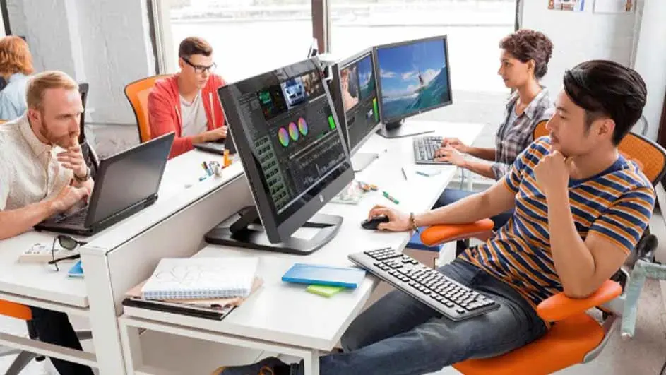 Modern office space with five colleagues working at computers. Bright workspace featuring orange chairs and multiple monitors for collaborative work.