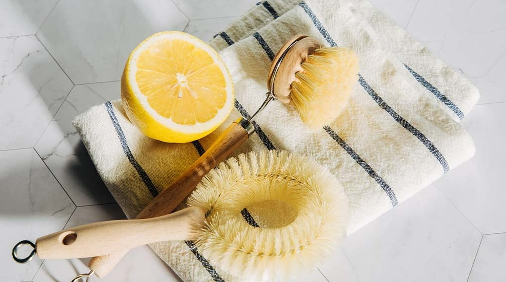 Un citron coupé, des brosses en bois et une serviette rayée sur un fond carrelé blanc.