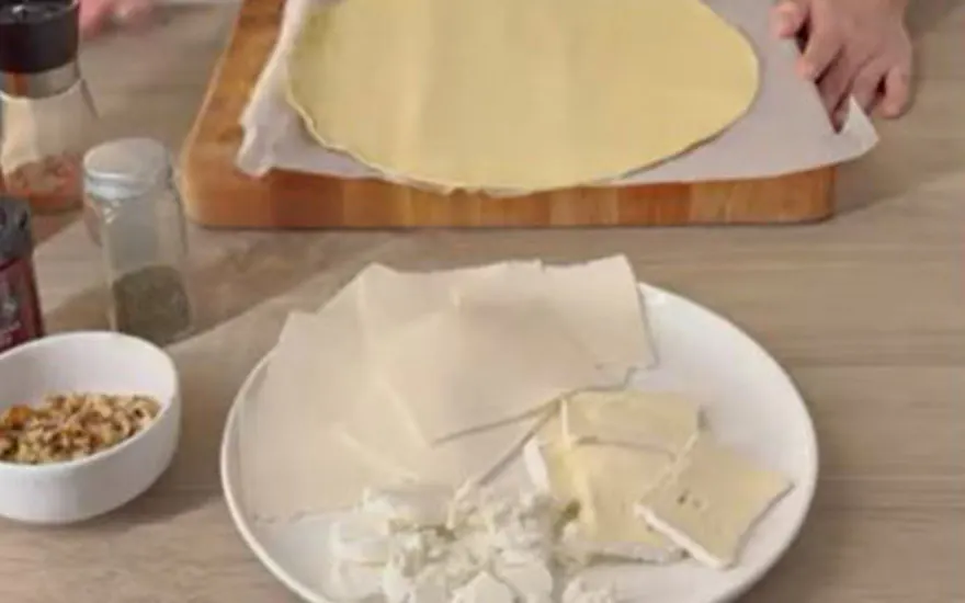 Une table présente une pâte ronde sur une planche, une assiette avec des feuilles, du fromage et un bol de noix.