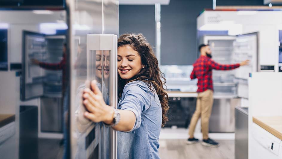 A showroom displays stainless steel refrigerators. A woman touches a fridge while another person examines a fridge in the background.
