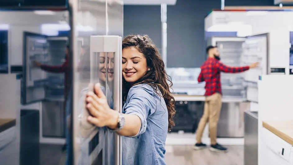 A showroom displays stainless steel refrigerators. A woman touches a fridge while another person examines a fridge in the background.