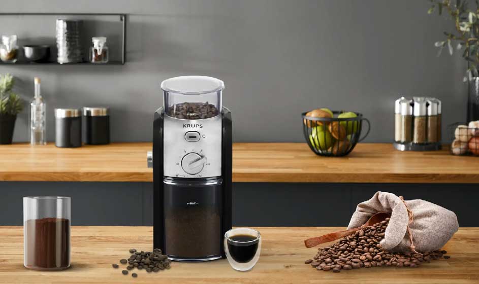 Een zwarte koffiemolen staat op een houten werkblad. Ernaast liggen koffiebonen, gemalen koffie en een kopje; achteraan staan potten en fruit.
