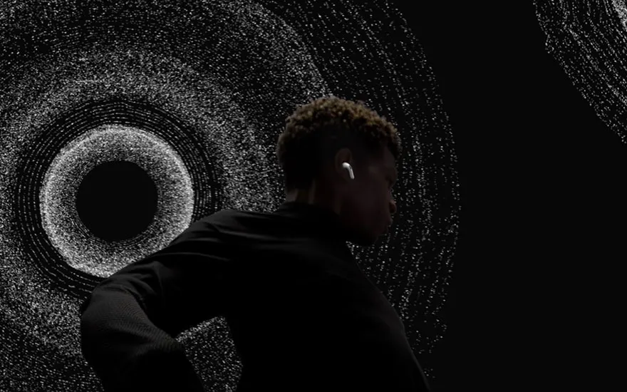 Un homme portant un écouteur sans fil, devant un fond noir avec des motifs circulaires gris et blancs.