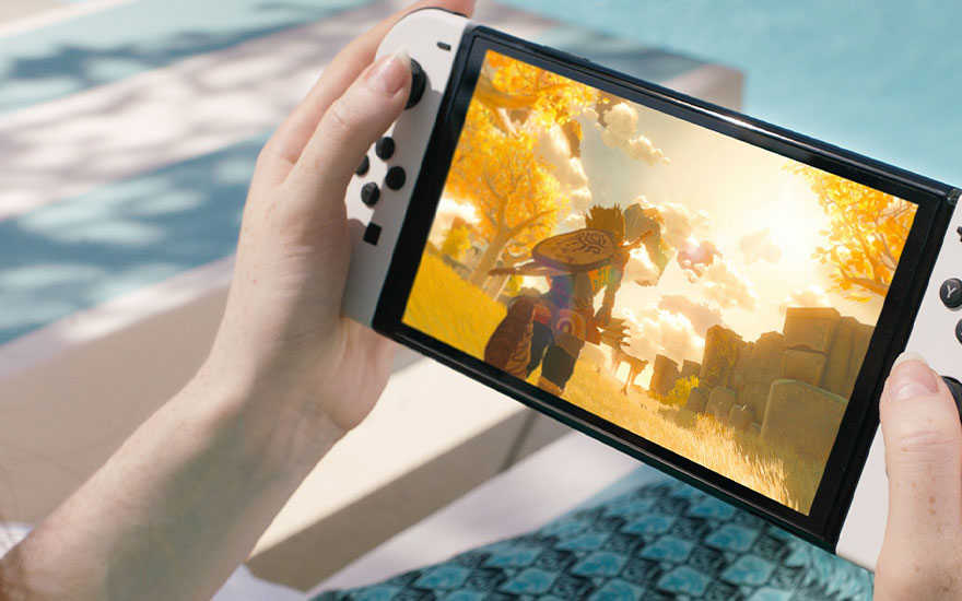 Un Nintendo Switch OLED blanc est tenu devant une piscine, affichant un jeu avec des arbres jaunes.