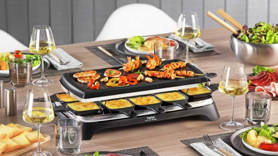 Een raclette-apparaat op een tafel met gegrilde ingrediënten, kaas, glazen wijn en salades.