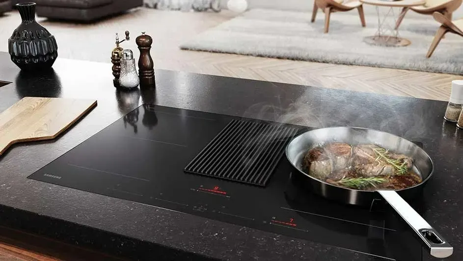 Cuisine sur plan noir avec une poêle en acier fumante. Ustensiles, planche à découper et salon en arrière-plan.