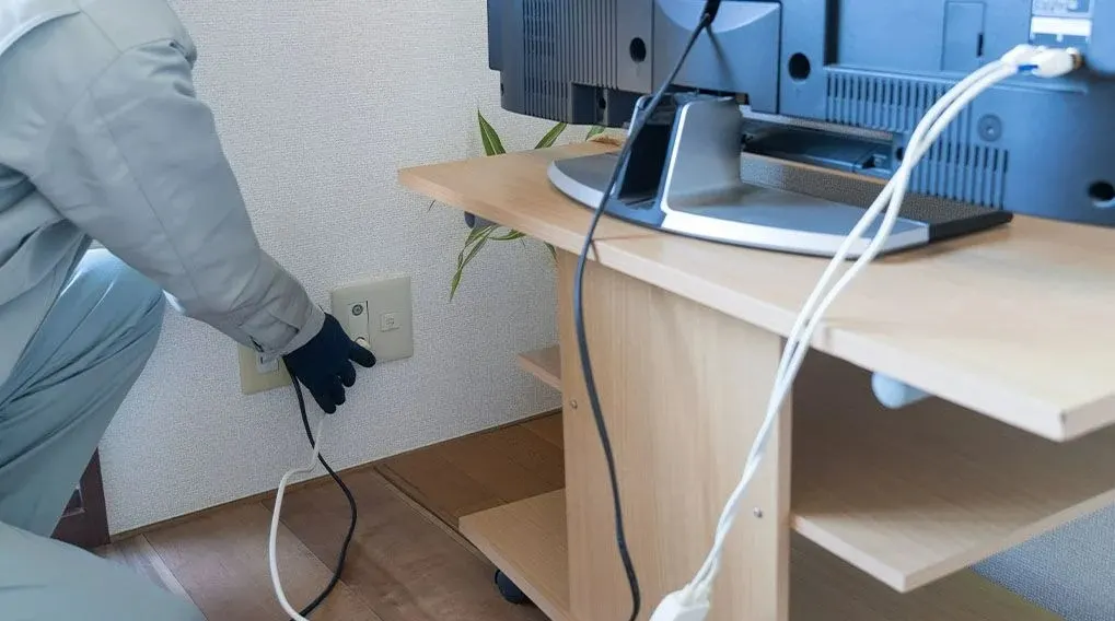 Un appareil TV est posé sur une table en bois claire. Une main gantée manipule une prise électrique murale blanche. Des câbles blancs et noirs sont visibles.